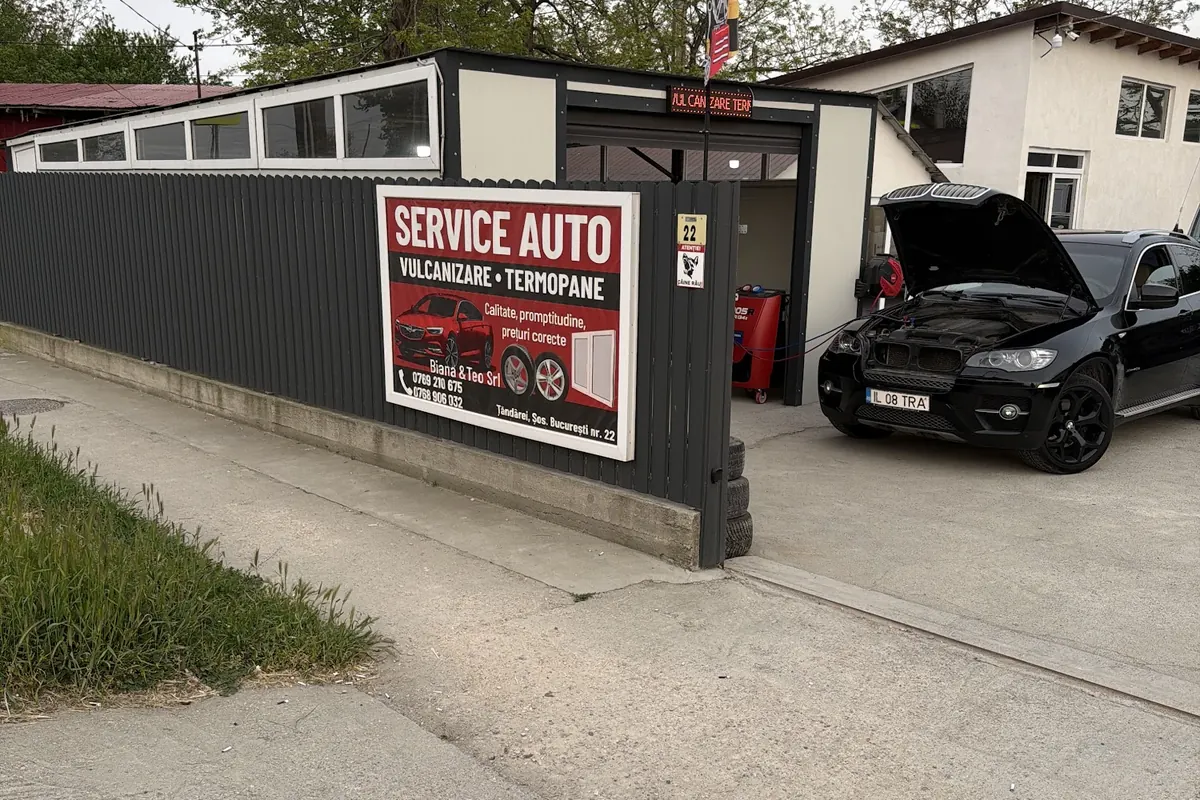 Gardul gri al service-ului, cu panoul roșu Service auto, numărul 22 la poartă și curtea în care se lucrează